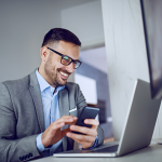 Smiling office worker holding mobile phone and looking at laptop